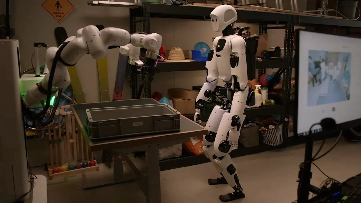 The Apollo 2 humanoid standing beside a bi-arm Franka robot at a workbench with a grey tote, a monitor at right showing the robot's camera view, as taken from Google DeepMind